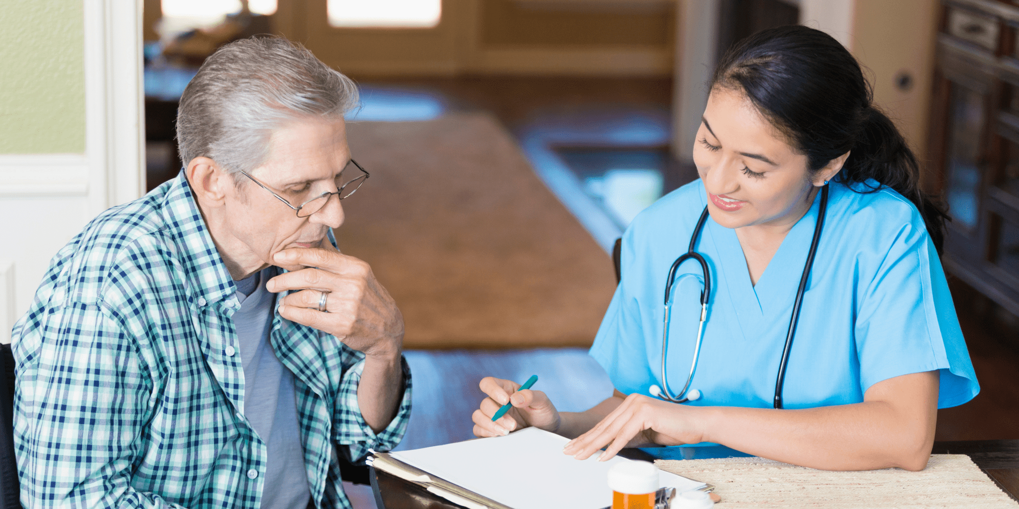 a home healthcare worker wearing scrubs and a stethoscope, while talking to an elderly man about his medication.